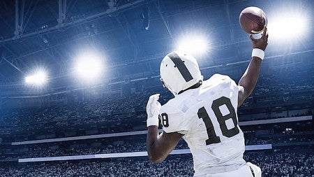 A football quaterback dressed in white with a white helmet throws a pass with the brighlights from a night game shining down
