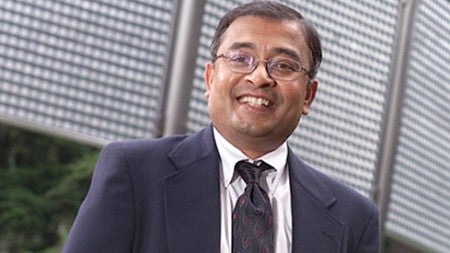 Photo of Nagesh Murthy in a suit standing in front of the wind fence art work on the University of Oregon campus