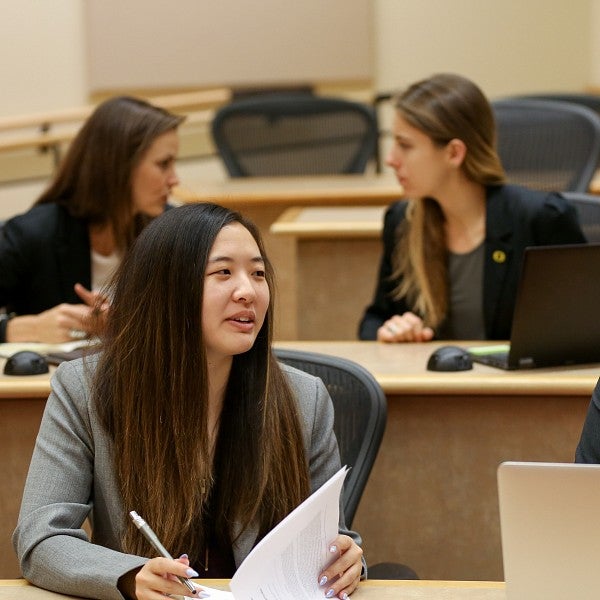 Four students sitting in a classroom with laptops and notebooks talking amongst themselves.